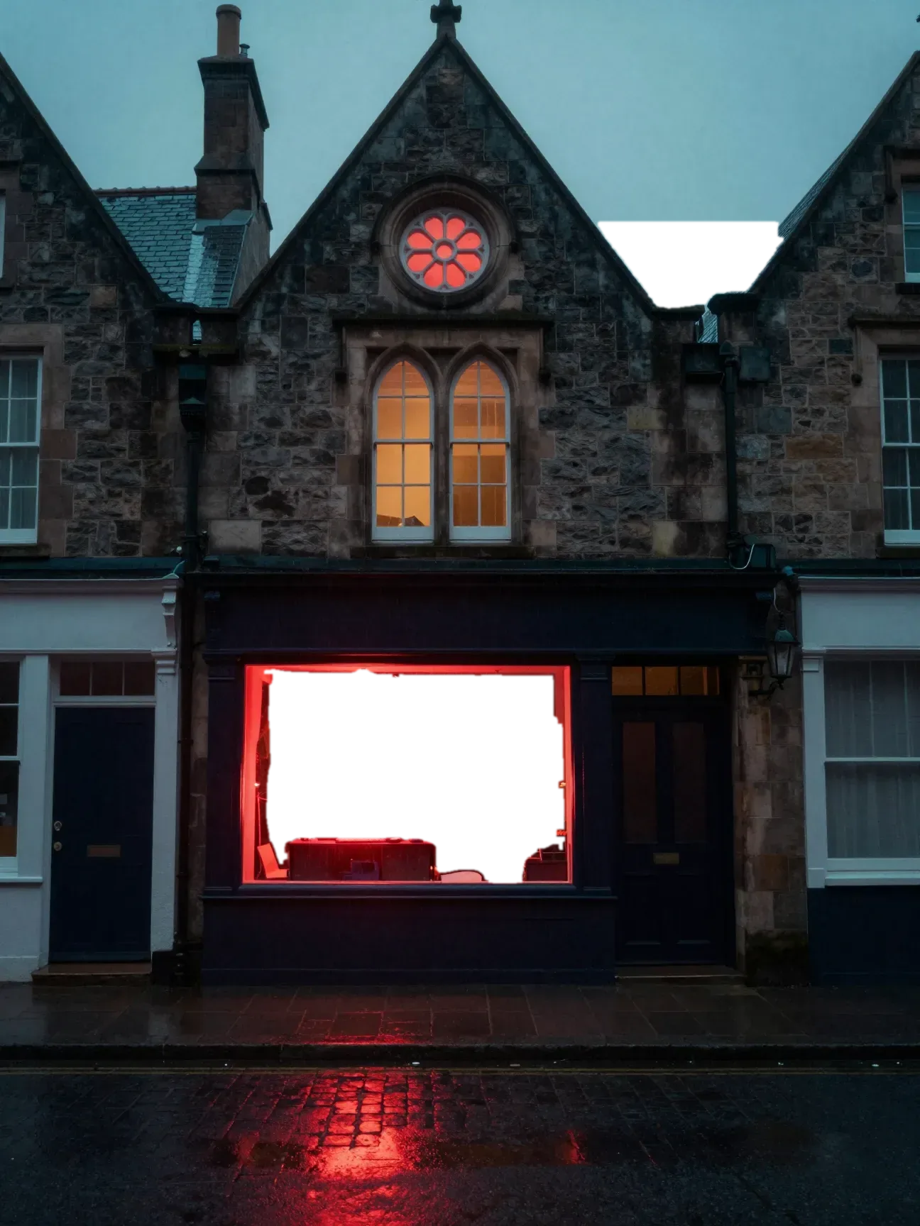 A gothic stone row house at night with a red rose window. Behind the shop glass, a computer monitor glows harsh red in a dark room.